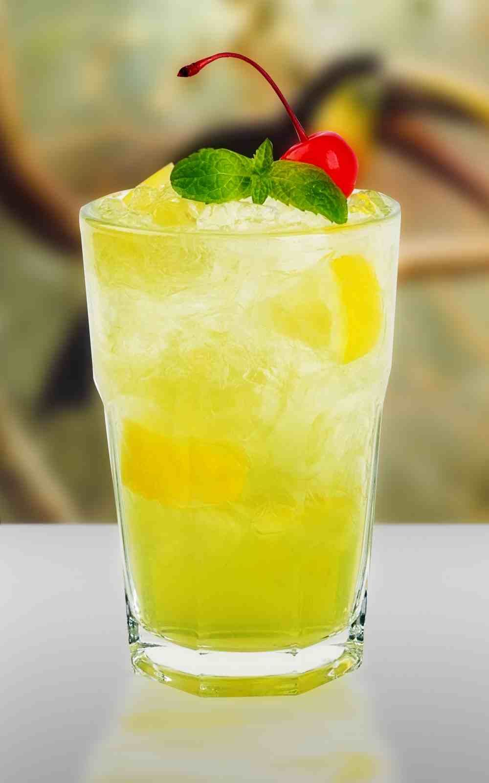 Szklanka z zielonym napojem, kostkami lodu, plasterkami cytryny, udekorowana wiśnią koktajlową i listkami mięty.